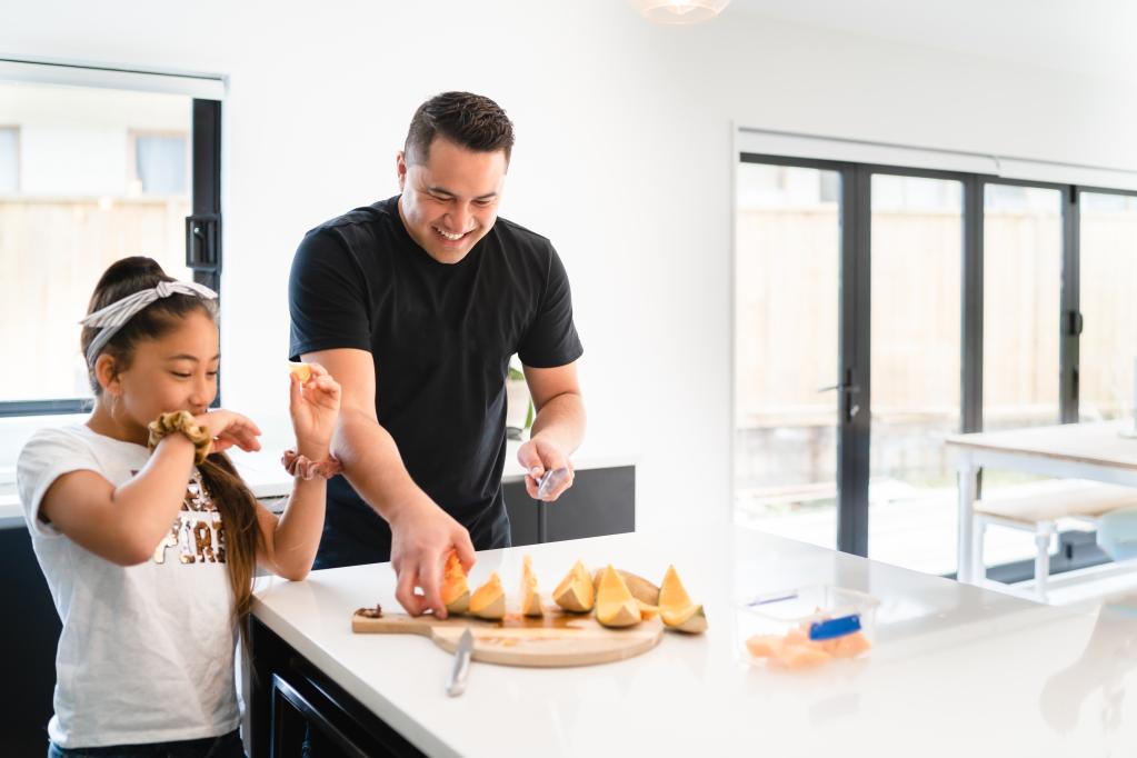 Adult and child preparing food together in a kitchen. The adult is cutting melon while the child eats a piece of fruit, showing a positive family mealtime and healthy eating.