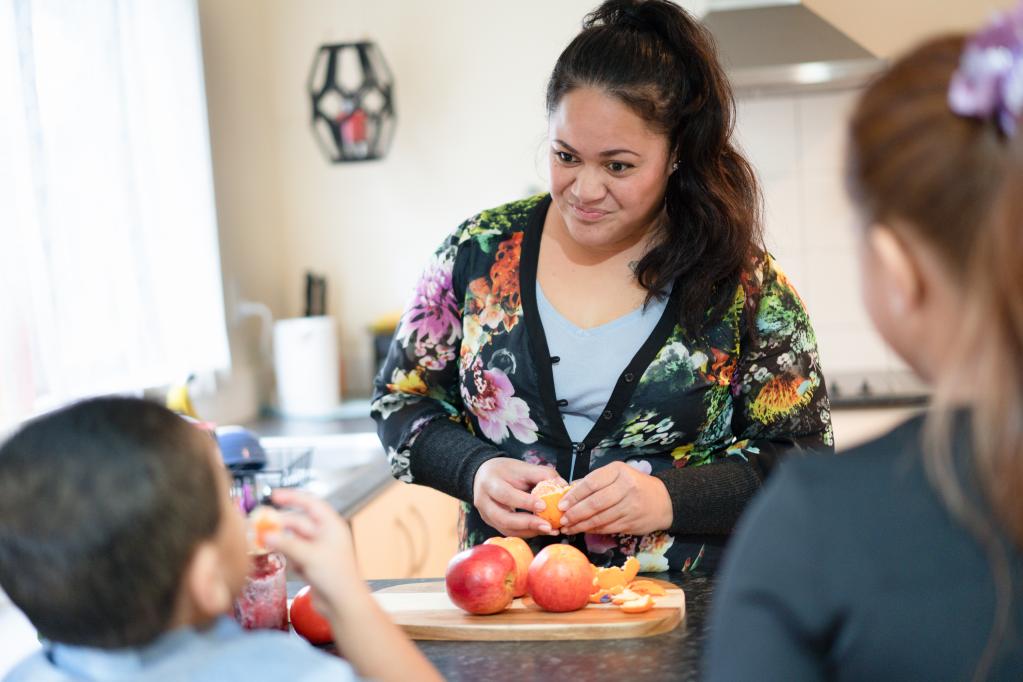 Parent preparing fresh fruit in the kitchen while 2 children watch and eat fruit together at the bench, showing a positive family mealtime and healthy eating.