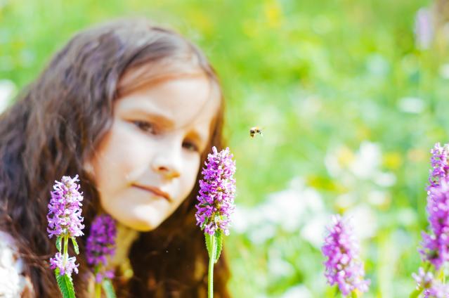 A young girl with long, wavy brown hair gazes at a bee hovering near a purple wildflower in a meadow. The scene is bright and filled with greenery, with additional wildflowers blurred in the background, giving a sense of depth and natural beauty. The girl’s expression is focused and curious as she observes the bee in close proximity."