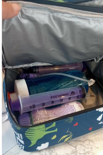 An open insulated cooler bag containing tube feeding supplies, including syringes, a feeding bottle, and medical equipment.