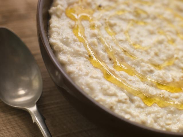 A close-up of a bowl of creamy porridge drizzled with honey, beside a metal spoon on a wooden table.