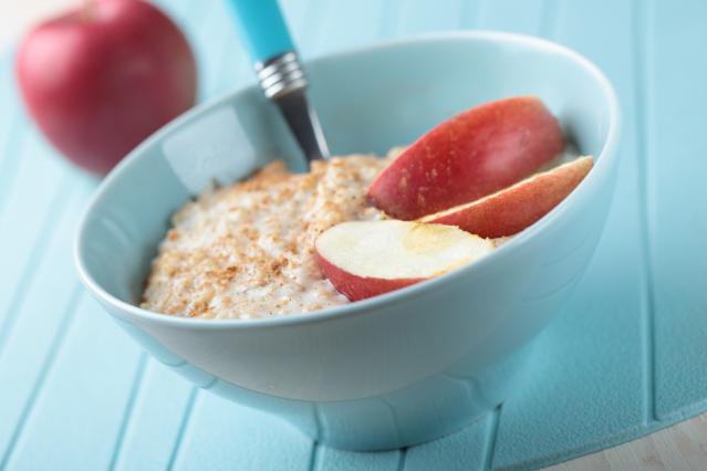 A bowl of creamy porridge topped with sliced red apple and cinnamon, with a spoon in the bowl and a whole apple blurred in the background.