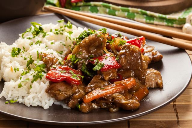 A plate of white rice served with pork and vegetable stir-fry in a glossy sauce, including red capsicum, carrot, mushrooms, and onion, garnished with chopped herbs.
