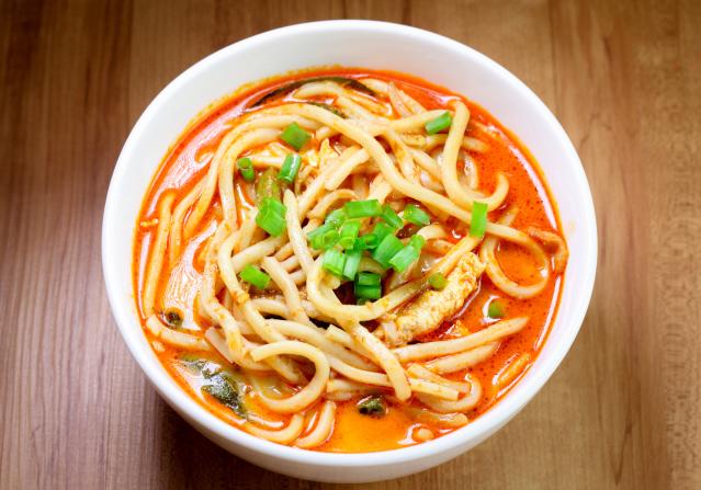 A bowl of noodle soup in a red-orange broth, topped with sliced spring onion, on a wooden table.