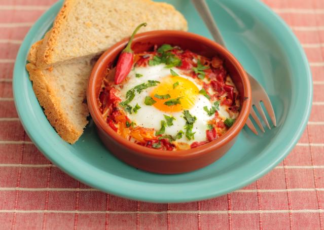 A baked egg dish with tomato and herbs served in a small brown ramekin on a blue plate, alongside slices of toasted bread and a fork.