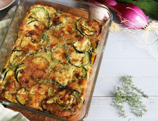 A baked zucchini and rice dish with melted cheese and herbs in a glass baking dish, surrounded by fresh zucchini, red onions, and thyme.