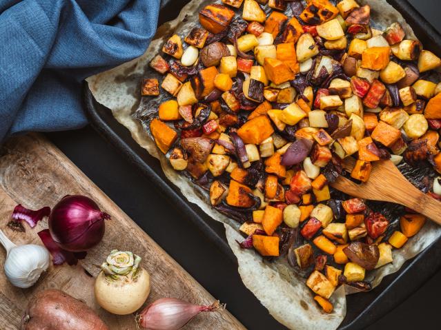A tray of roasted vegetables, including kūmara, potato, carrot, onion, and garlic, on baking paper with a wooden spoon, beside fresh vegetables on a wooden board.