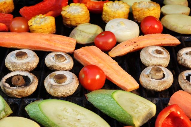 A variety of vegetables cooking on a barbecue grill, including mushrooms, zucchini, kumara, corn, onion, cherry tomatoes, and red capsicum.