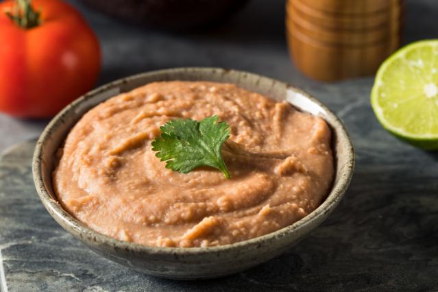 Smooth bean dip in a bowl topped with a coriander leaf, with tomato and lime in the background.