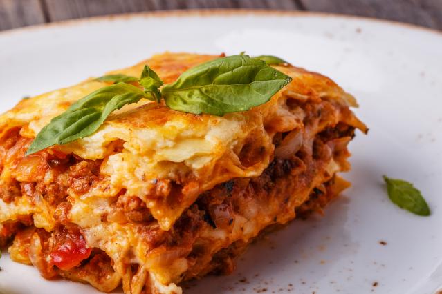 A slice of beef lasagne with melted cheese and layers of pasta and meat sauce, garnished with fresh basil leaves on a white plate.