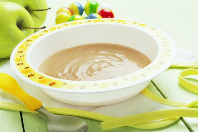 A bowl of smooth fruit purée beside a baby spoon, with green apples and colourful toy balls in the background.