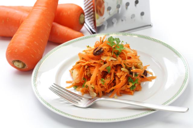 A carrot and orange salad with raisins and nuts on a white plate, with whole carrots and a grater in the background.