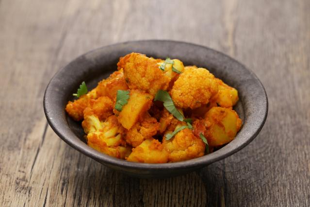 Bowl of cauliflower and potato curry garnished with fresh herbs on a wooden table.