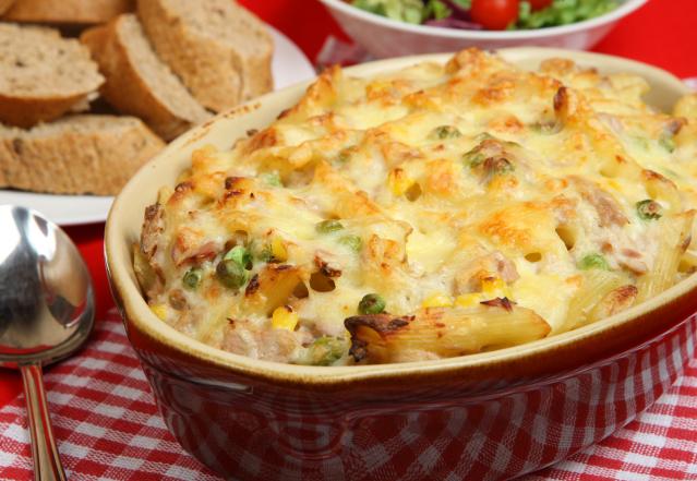 A baked chicken and vegetable pasta dish topped with melted cheese in a casserole dish, with bread and salad in the background.