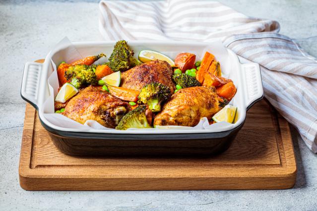 Roasted chicken thighs with broccoli, carrots, and lemon wedges served in a baking dish on a wooden board.