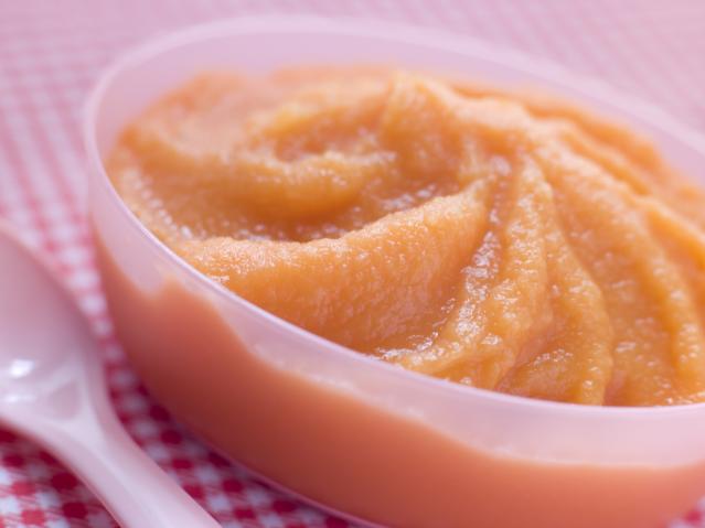A bowl of smooth chickpea and kūmara purée beside a baby spoon on a pink checkered cloth.