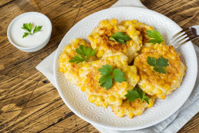 A plate of golden corn fritters topped with parsley leaves, served with a small bowl of white yoghurt or sour cream dip.