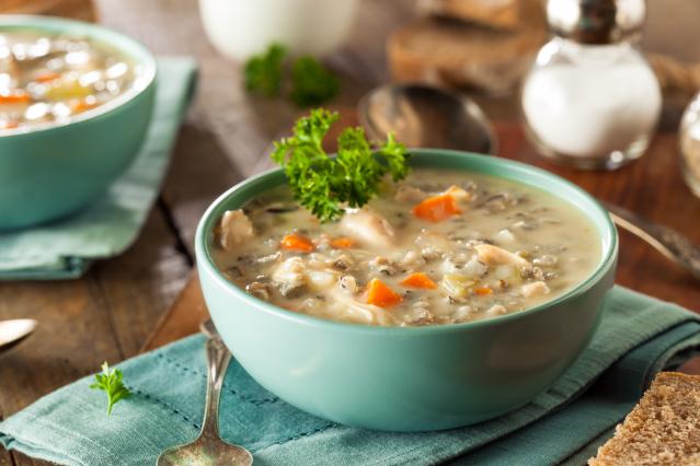 A bowl of creamy chicken and vegetable soup with carrot and parsley, served on a wooden table with bread nearby.