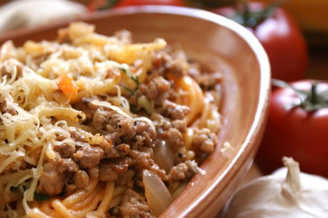 Bowl of curried mince with vegetables served on spaghetti and topped with grated cheese, with tomatoes and garlic blurred in the background.