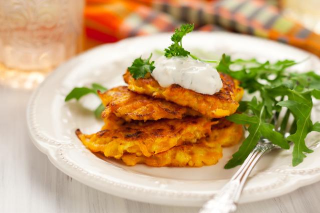 Stack of golden vegetable fritters topped with a creamy herb sauce and parsley, served on a white plate with rocket leaves and a fork, with a softly blurred table setting in the background.