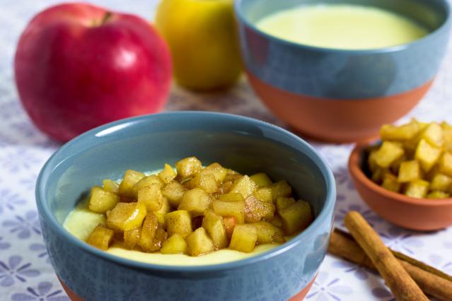 Bowls of creamy custard topped with diced cooked apple and cinnamon, with fresh apples and cinnamon sticks in the background.