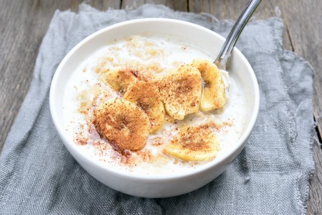 Bowl of creamy porridge topped with sliced banana and a sprinkle of cinnamon, served with a spoon on a grey cloth over a rustic wooden table.