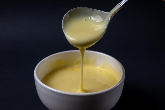 Spoon lifting thick creamy custard above a white bowl, with the custard slowly dripping back into the bowl against a dark background.