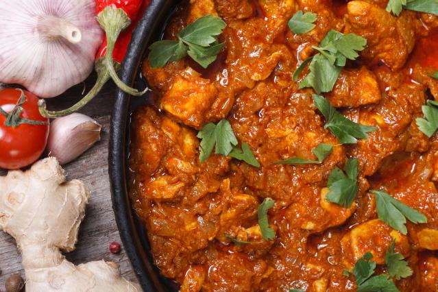 Close-up of a rich butter chicken curry garnished with fresh parsley in a black serving dish, with garlic, ginger, tomatoes and red chilli arranged on a rustic wooden table beside the dish.