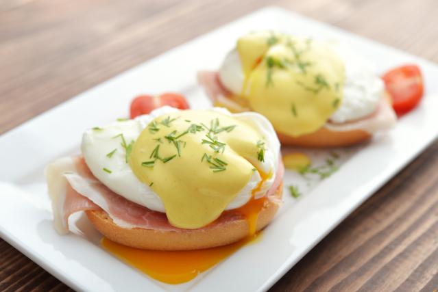 Two eggs Benedict served on toasted English muffins with ham, poached eggs and creamy hollandaise sauce, garnished with herbs on a white plate on a wooden table.