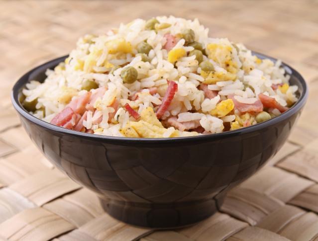 Bowl of fried rice with peas, egg and sliced ham, served in a black bowl on a woven placemat background.