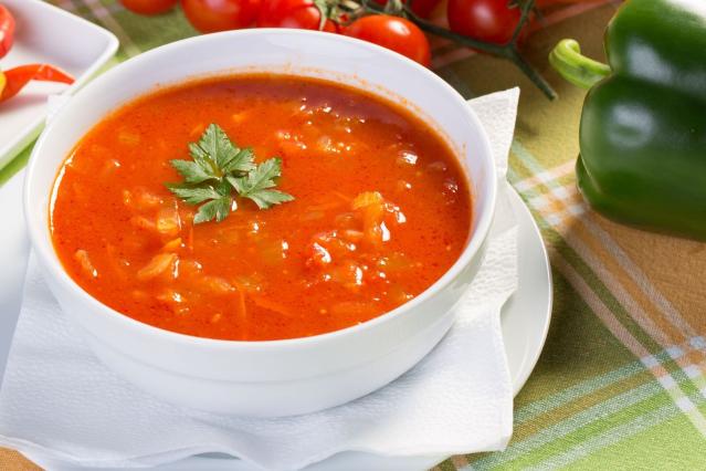 Bowl of tomato-based vegetable soup garnished with parsley, served on a plate with fresh tomatoes, chillies and capsicums arranged on a checked tablecloth in the background.