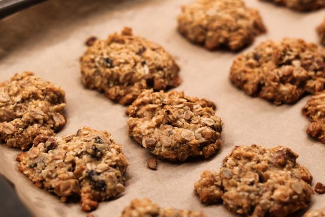 Freshly baked oat cookies with visible oats and dried fruit cooling on a baking tray lined with baking paper, photographed close up.