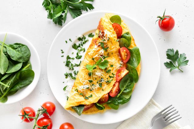ALT text: A folded omelette filled with spinach and cherry tomatoes on a white plate, topped with grated cheese and chopped herbs. The plate sits on a light table surrounded by fresh spinach leaves, parsley, cherry tomatoes, and a fork.