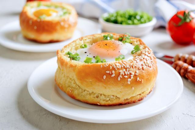 A baked egg served inside a sesame seed bread roll on a white plate, topped with chopped spring onion and black pepper. Another egg-filled bread roll, fresh herbs, tomatoes, and nuts are blurred in the background on a light table setting.