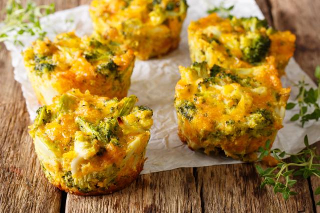 Five baked egg muffins with broccoli and melted cheese sitting on baking paper on a rustic wooden table, with small sprigs of fresh herbs scattered around them.