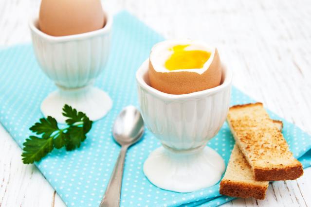 Two soft-boiled eggs in white egg cups on a light wooden table, with one egg cracked open to show a runny yolk. Toast soldiers, a teaspoon, and parsley sit beside the eggs on a blue polka dot napkin.