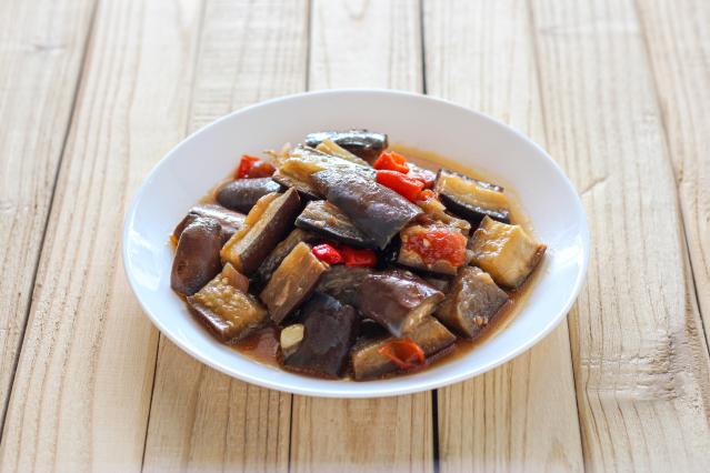 A white bowl filled with eggplant curry made with chunks of aubergine, red chilli, and sauce, sitting on a light wooden table.