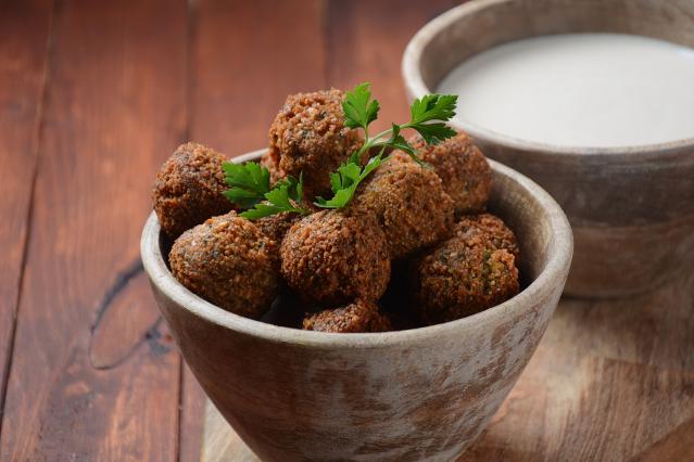 A wooden bowl filled with crispy falafel balls garnished with parsley, sitting on a rustic wooden table beside a small bowl of dipping sauce.