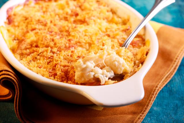 A baked fish pie with a golden crumb topping in a white oven dish, with a spoon scooping out the creamy filling. The dish sits on an orange cloth on a blue table surface.