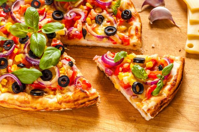 A colourful homemade pizza topped with black olives, red onion, capsicum, sweetcorn, ham, cheese, and fresh basil on a wooden board, with one slice separated from the rest. Garlic cloves and slices of cheese sit nearby.
