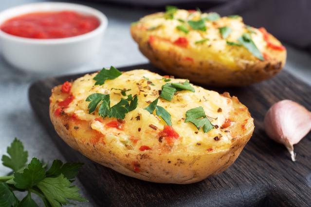 Two baked potato halves filled with melted cheese, herbs, and small pieces of red capsicum sit on a dark wooden serving board. Fresh parsley, a garlic clove, and a small bowl of tomato sauce are arranged nearby on a kitchen bench.