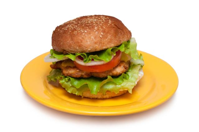 A fish burger with a sesame seed bun sits on a bright yellow plate against a plain light background. The burger is filled with a crumbed fish fillet, lettuce, tomato, onion, and melted cheese.