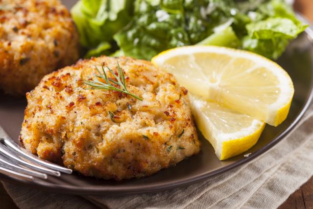 Two golden-brown fish cakes are served on a dark plate with fresh lettuce leaves and lemon wedges. A sprig of dill sits on top of the fish cake in the foreground. The plate is set on a wooden table with a beige cloth napkin and a fork nearby.