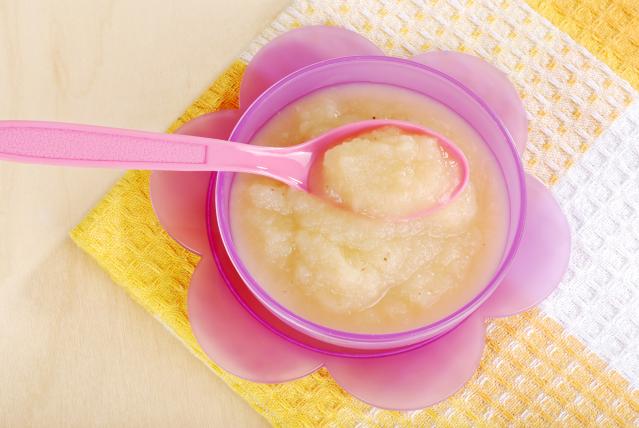 A pink spoon rests in a pink flower-shaped bowl filled with smooth pale fish purée. The bowl sits on a yellow and white textured cloth on a light wooden table.
