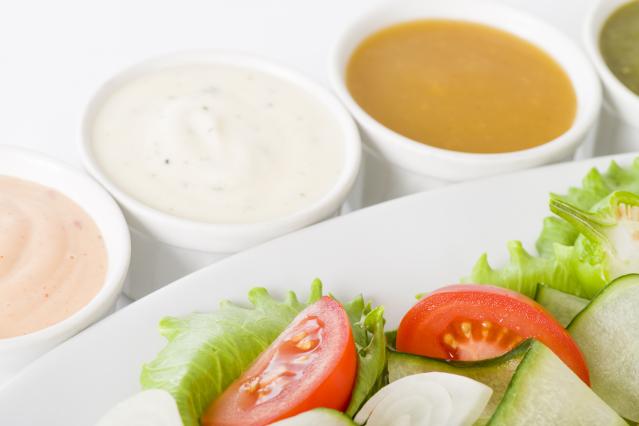 Small white bowls of salad dressings, including creamy white and orange dressings, sit beside a fresh salad with lettuce, tomato, cucumber, and onion on a white plate against a plain light background.