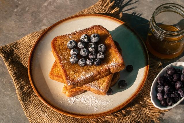 A stack of golden French toast dusted with icing sugar and topped with fresh blueberries sits on a ceramic plate. Nearby are a jar of honey and a small bowl of blueberries on a rustic woven cloth over a grey tabletop.