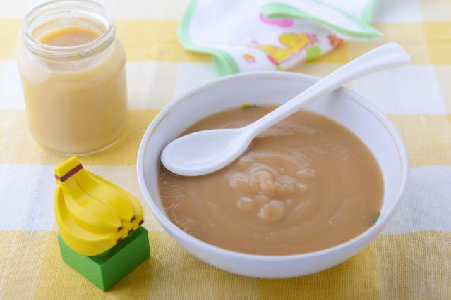 A bowl of smooth fruit purée with a white spoon sits on a yellow checked cloth beside a small jar of purée. A toy banana block and folded baby bib are nearby on the table.