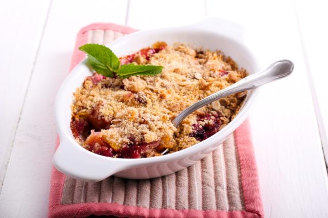 A baked fruit crumble with a golden crumb topping sits in a white ceramic dish on a pink quilted mat. A spoon rests in the crumble, and a sprig of fresh mint sits on top. The dish is placed on a white wooden table.