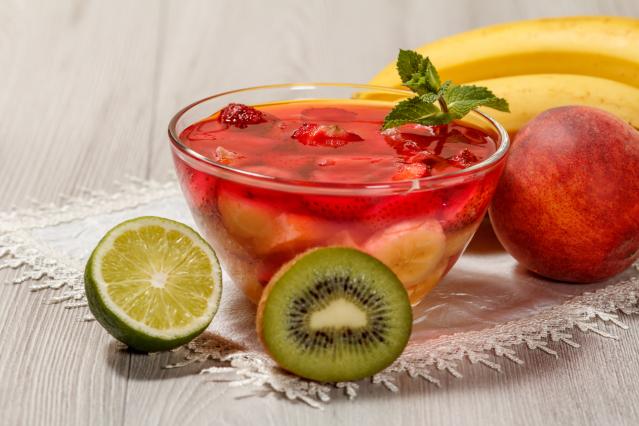 A glass bowl of red fruit jelly with pieces of banana and strawberry sits on a lace cloth on a wooden table. Fresh fruit including lime, kiwi fruit, bananas, and a peach are arranged around the bowl, with a sprig of mint on top of the jelly.