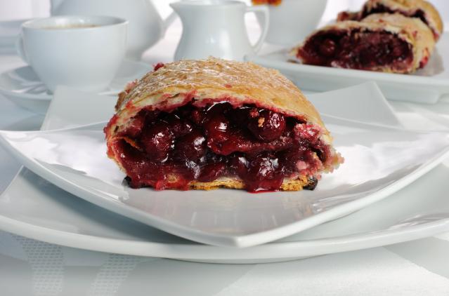 A slice of berry-filled strudel with flaky pastry sits on a white plate. Dark red berry filling is visible inside the pastry, with another piece of strudel and white cups and jugs blurred in the background.